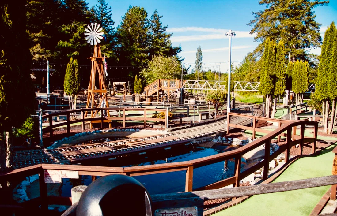 The Station 49 mini golf course — landscaped greens with the windmill, pond, footbridge, and the train track running through